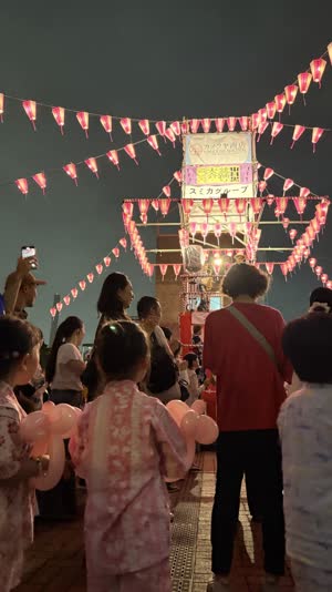 High overhead view of a large circle of dancers surrounding a lantern-lit yagura tower at night, concentric ring formation, warm lights against dark ground, people too distant…