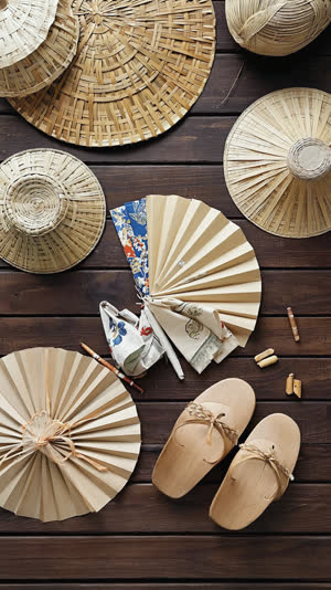 Overhead flat lay of festival dance items: woven straw hats, folded tenugui cloths, paper fans and wooden geta sandals on dark wood, varied regional textures, soft even light