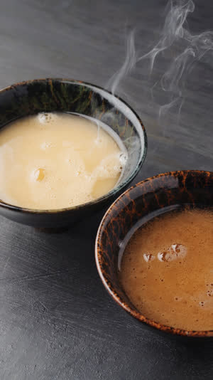 Overhead shot of two lacquer bowls of miso soup side by side, one pale creamy gold and one deep dark brown, steam rising, dark wooden table, soft natural light