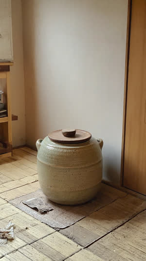 Old glazed ceramic crock with a wooden lid sitting in the corner of a lived-in Japanese kitchen, soft morning light through a window, quiet domestic mood