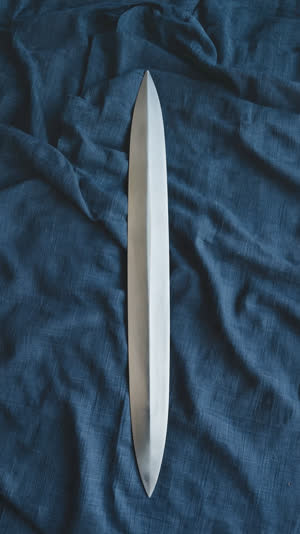 Single unfinished sword blade resting on dark indigo cloth, vast negative space, overhead composition, matte raw steel texture, contemplative minimalism
