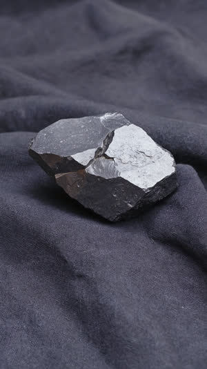 A rough chunk of raw tamahagane steel on dark cloth, dull grey iron with a matte rough surface, only the fractured face showing a subtle silvery crystalline sheen, photorealistic…