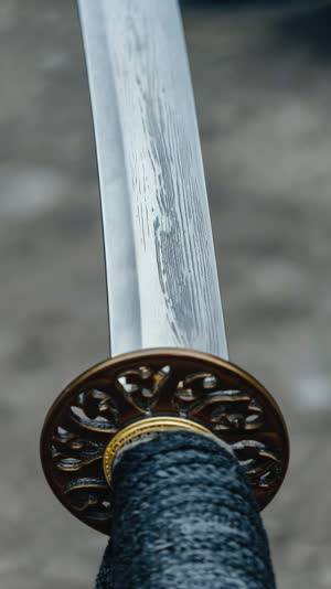 Shallow-angle close-up of the middle section of a polished Japanese katana blade, both ends out of frame, cool silver-grey metal with a faint subtle layered banding in the steel…
