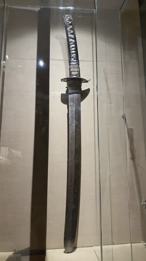 Katana blade behind museum glass viewed up close, wood-grain jihada texture visible along the steel, focused gallery spotlights, dark hall