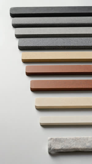Overhead view of about ten Japanese whetstones arranged from coarse to ultra-fine, varied earthy colors and sizes, tiny finishing stone slivers at the end of the row, neutral…