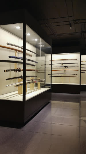 Japanese sword museum gallery, blades displayed in glass cases under focused spotlights, hamon patterns clearly visible, dark elegant exhibition room
