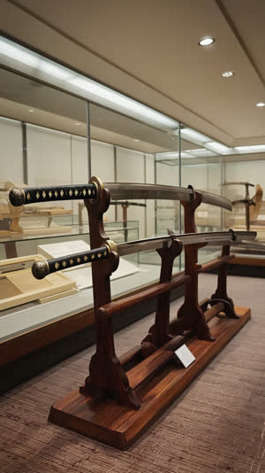 Museum display of historic Bizen swords on wooden stands, blades in sharp focus under soft exhibition lighting, background exhibits as gentle bokeh, no documents, no labels, no…