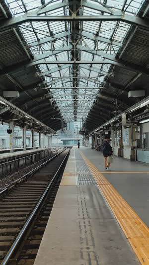 Wide view of a Japanese train platform, rhythmic lines of tracks, painted marks and roof beams, distant blurred figures