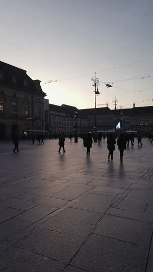 Calm station square at dusk, long shadows and an orderly flow of distant silhouettes, muted respectful tones, wide shot