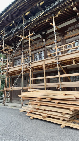 Scaffolding and stacked replacement timbers beside a Japanese temple building under restoration, work in progress, photorealistic documentary photograph, natural soft light,…