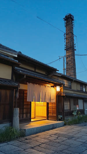 Traditional Japanese public bathhouse exterior at dusk, glowing entrance with fabric noren curtain, tall chimney against twilight sky, warm light spilling out, nostalgic film…