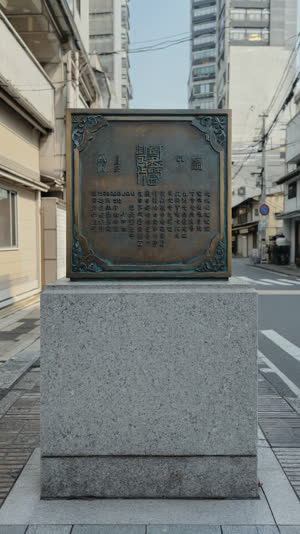 Commemorative metal plaque on a stone base at a quiet Tokyo street corner, engraved Japanese text, urban Kanda backdrop softly blurred, documentary style