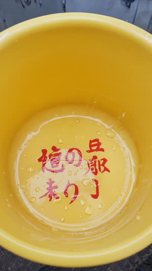 Overhead close-up of yellow bath bucket interior with red Japanese lettering printed on the bottom, water droplets on plastic, crisp graphic detail