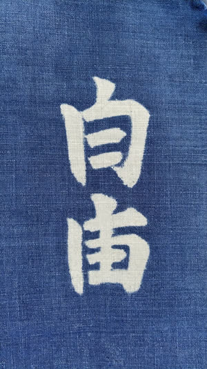 Close-up of white dyed Japanese hiragana character on indigo fabric noren, visible weave texture and dye edges, soft natural light, tactile detail