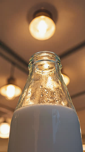 Dramatic low-angle shot of a glass milk bottle tilted upward against retro ceiling lights, heroic composition, condensation on glass, nostalgic tones