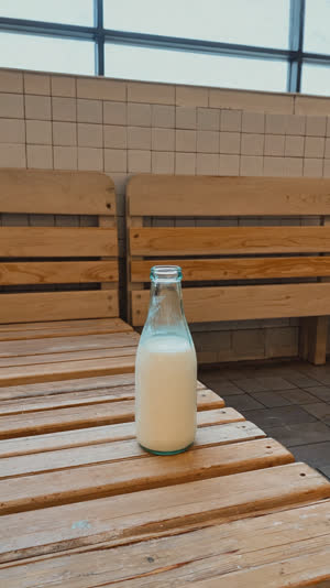 Empty glass milk bottle resting on wooden bench in quiet bathhouse changing room, soft evening light, peaceful afterglow atmosphere