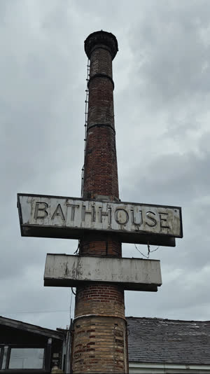Looking up at a faded bathhouse sign and old chimney against overcast sky, weathered lettering, solemn documentary tone, muted colors