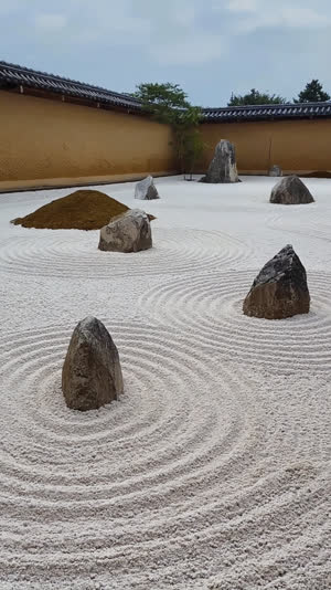 A Zen dry landscape garden with groups of natural angular weathered rocks set in raked white gravel, low earthen wall, quiet overcast light, natural irregular weathered rocks…
