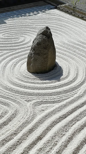 One rugged natural weathered rock rising from raked white gravel with concentric ripple lines around it like an island in a current, evocative close-up, natural irregular…