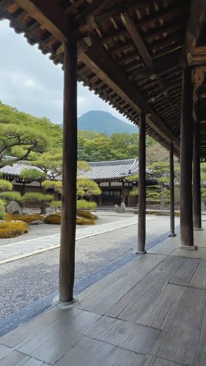 View from inside a Japanese temple veranda: dark wooden pillars and deep eaves framing a garden and one distant blue-green peak like a painting, engawa architectural framing,…
