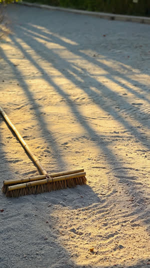 Early morning garden with long low light, a bamboo broom leaning against a stone, freshly swept gravel lines, photorealistic documentary photograph, natural soft light, muted…