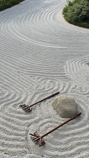 Close-up of long curved parallel rake lines in white gravel sweeping around the base of one natural weathered boulder in a full-size Japanese garden, realistic scale, discipline…