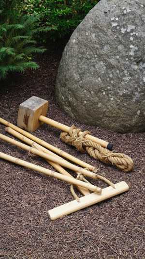 Traditional Japanese stone-setting tools resting on the ground beside a large natural garden boulder: a wooden mallet, coiled hemp rope, bamboo poles and a wooden lever,…