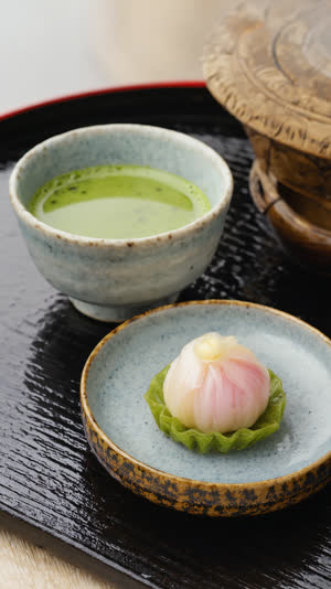 An elegant seasonal Japanese sweet beside a bowl of matcha on a lacquer tray, refined close-up, photorealistic documentary photograph, natural soft light, muted realistic colors,…