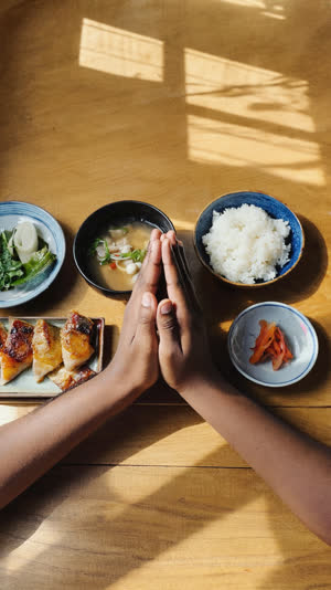 First-person point of view looking down at a Japanese breakfast set, the viewer's own two hands joined palm-to-palm in itadakimasu gesture, both forearms entering the frame from…