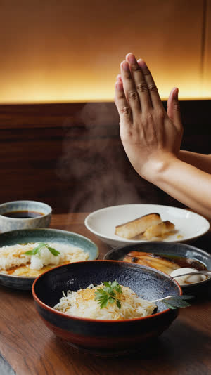 Hands beginning to press together over a steaming Japanese set meal, side view, face out of frame, warm intimate restaurant light