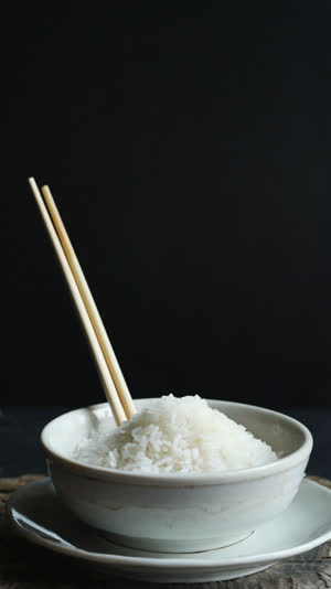 Chopsticks standing vertically in a mounded bowl of white rice, side angle, stark quiet lighting, dark backdrop, solemn atmosphere