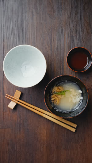 Overhead flat lay of a formal washoku place setting, rice bowl front-left, soup bowl front-right, chopsticks in front, clean documentary style