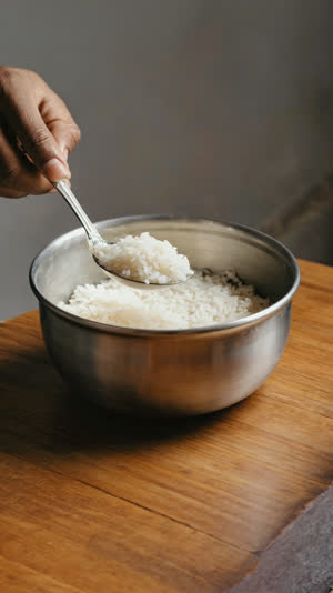 A hand scooping rice with a long Korean spoon while the stainless bowl stays on the table, close-up on hands, warm light
