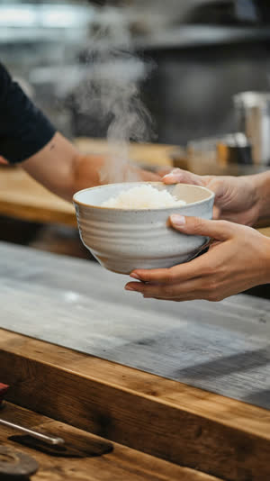 Hands lifting a rice bowl at a casual Japanese restaurant counter, steam rising, everyday warm atmosphere, blurred counter background