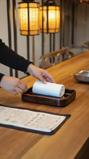 Hand offering a rolled steaming towel on a tray beside a closed menu on a wooden counter, warm izakaya light