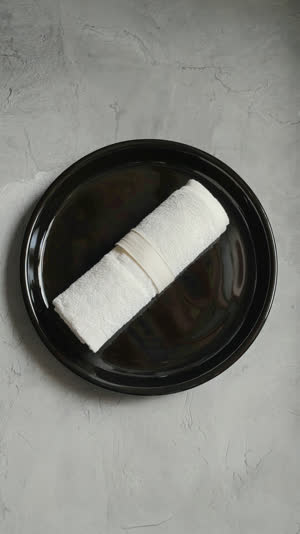 Single rolled white oshibori towel centered on a black lacquer tray, overhead flat lay, generous negative space, minimal composition, quiet directional light