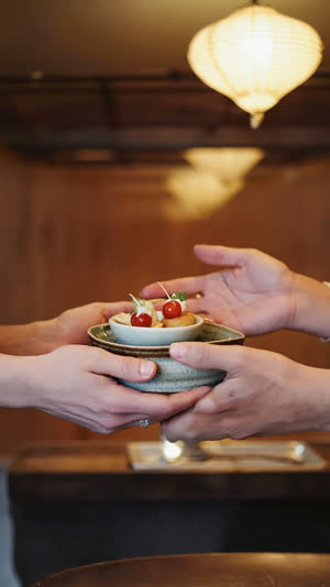 Two hands offering a small appetizer bowl forward with care, respectful serving gesture, warm izakaya glow