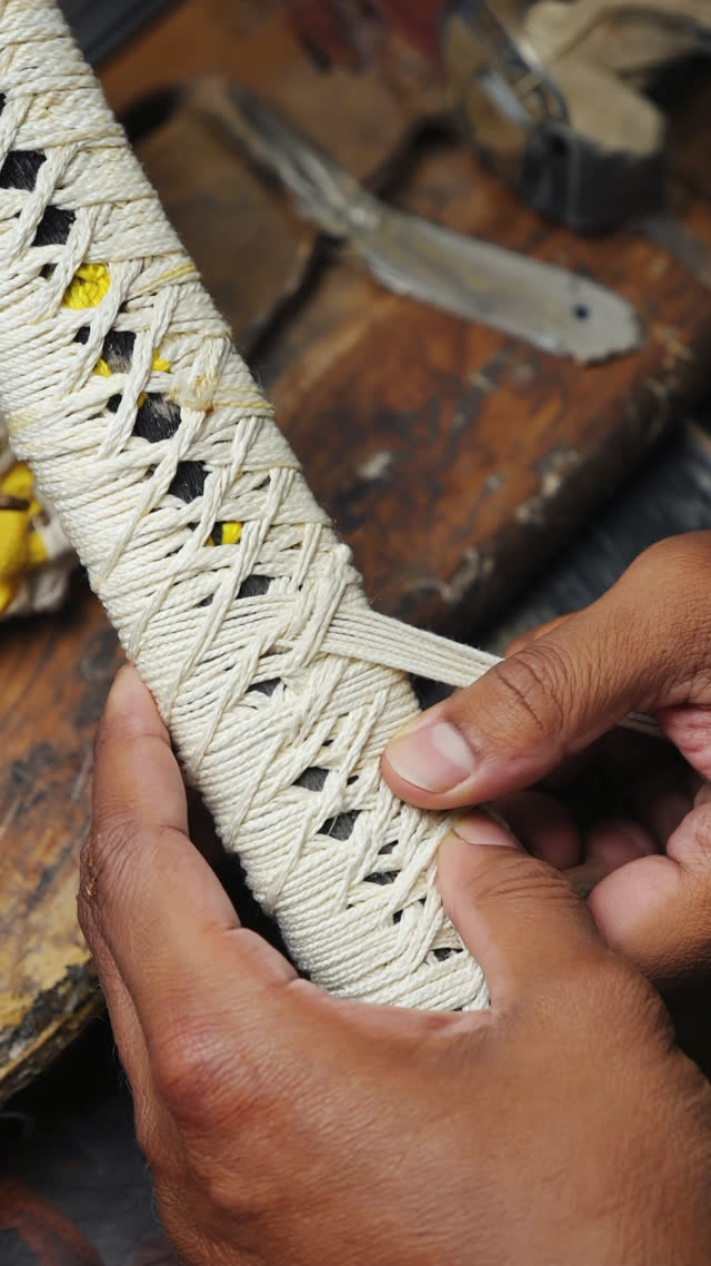 Close-up of hands wrapping silk cord in a precise diamond pattern around a Japanese sword hilt with white ray skin underneath, half-finished wrap showing the craft process,…