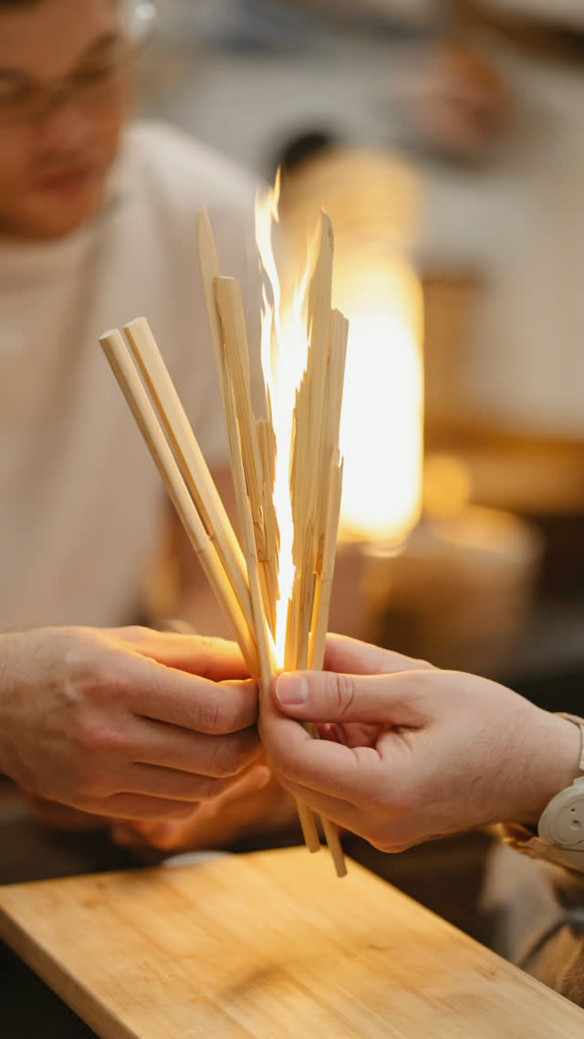 Hands holding freshly split cedar chopsticks, bright white split surfaces toward camera, close crop on hands, warm izakaya light