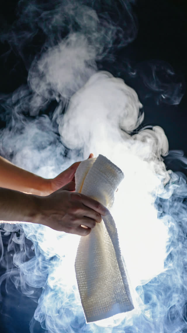 Macro of hands unrolling a hot white towel, steam rising and curling in backlight, fabric unfolding mid-motion, dark background