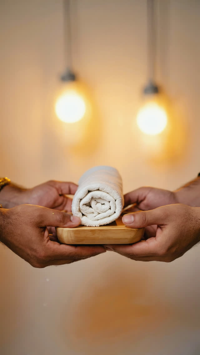 Two hands presenting a warm rolled towel forward on a small tray, respectful offering gesture, shallow depth of field, warm glow