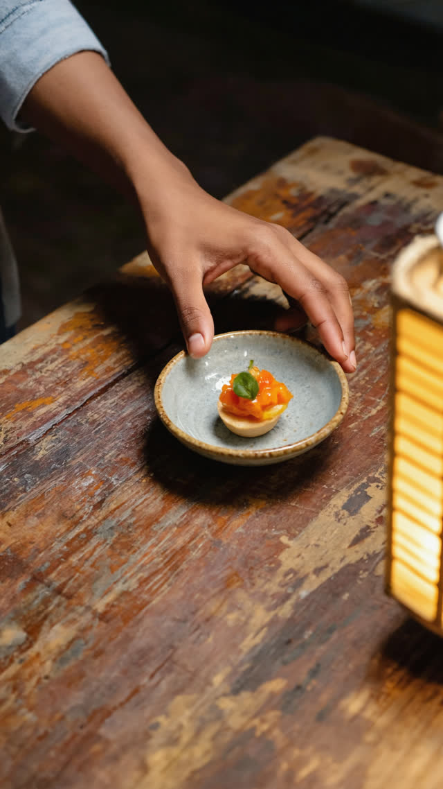 Hand setting down a small appetizer bowl on a wooden izakaya table, moment of placement, warm lantern light