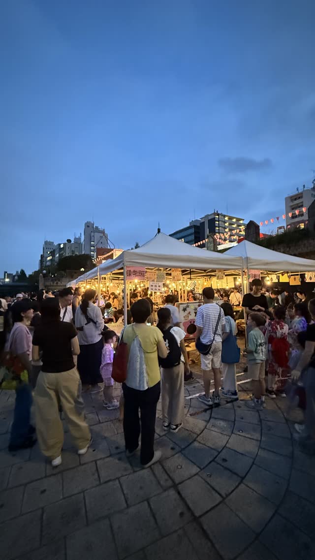 Wooden yagura tower in a town square at dusk, strings of glowing red paper lanterns radiating outward, festival about to begin, distant silhouettes only, warm evening light