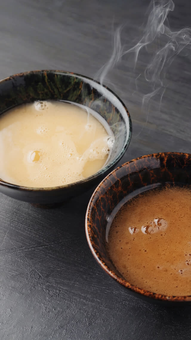 Overhead shot of two lacquer bowls of miso soup side by side, one pale creamy gold and one deep dark brown, steam rising, dark wooden table, soft natural light