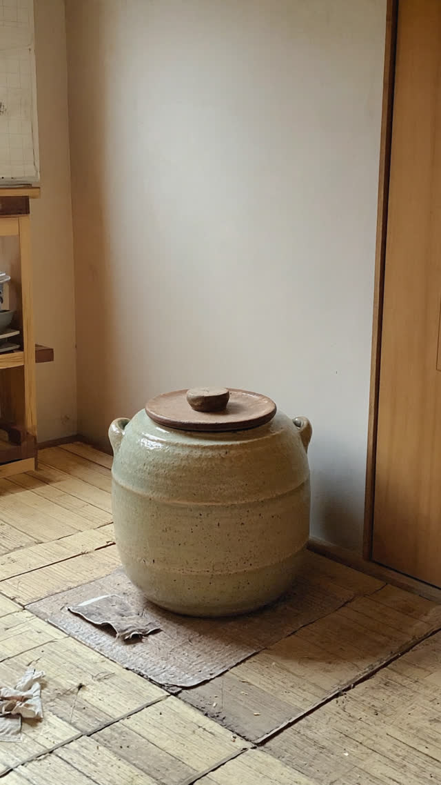 Old glazed ceramic crock with a wooden lid sitting in the corner of a lived-in Japanese kitchen, soft morning light through a window, quiet domestic mood