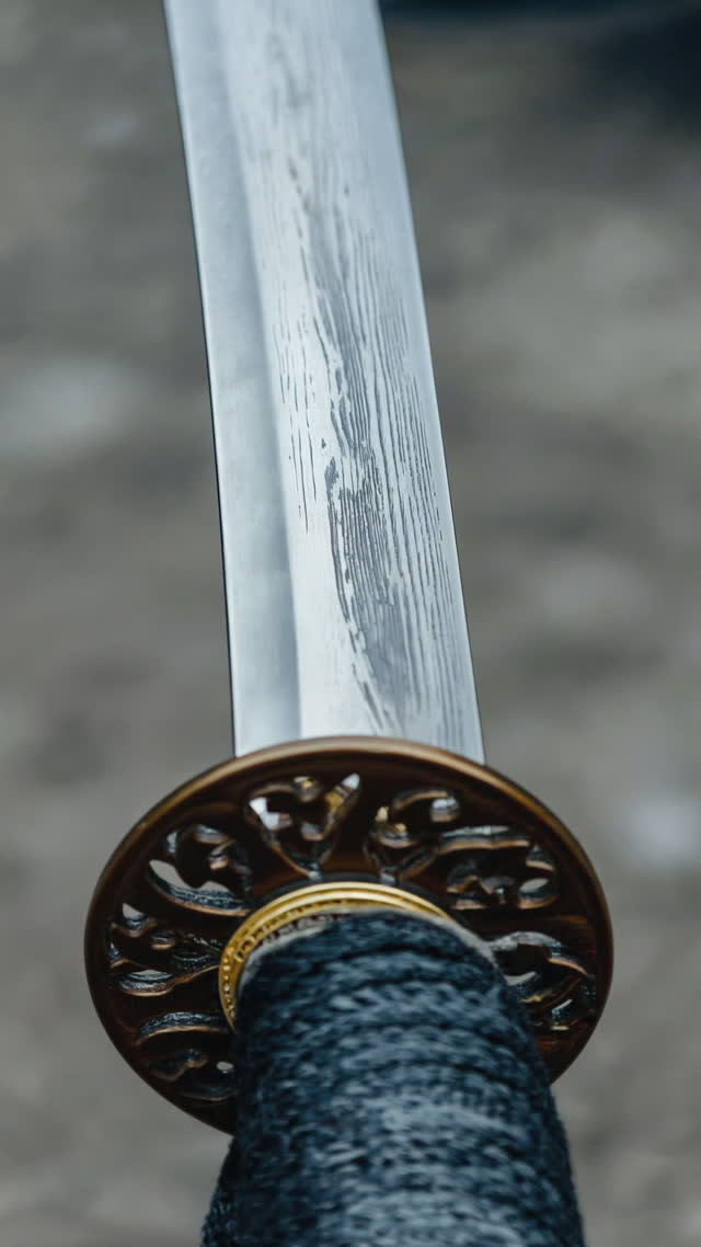 Shallow-angle close-up of the middle section of a polished Japanese katana blade, both ends out of frame, cool silver-grey metal with a faint subtle layered banding in the steel…