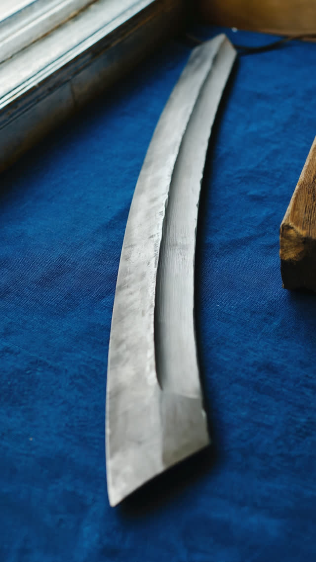 Close-up of the middle section of a freshly forged curved steel blade lying on indigo blue cloth, both ends out of frame, dull matte grey steel with rough forge scale texture and…