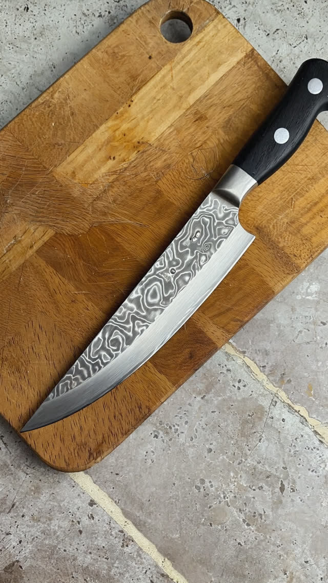 A Japanese santoku kitchen knife with a subtle hammered finish resting on a wooden cutting board in a home kitchen, soft daylight from a window, photorealistic documentary…