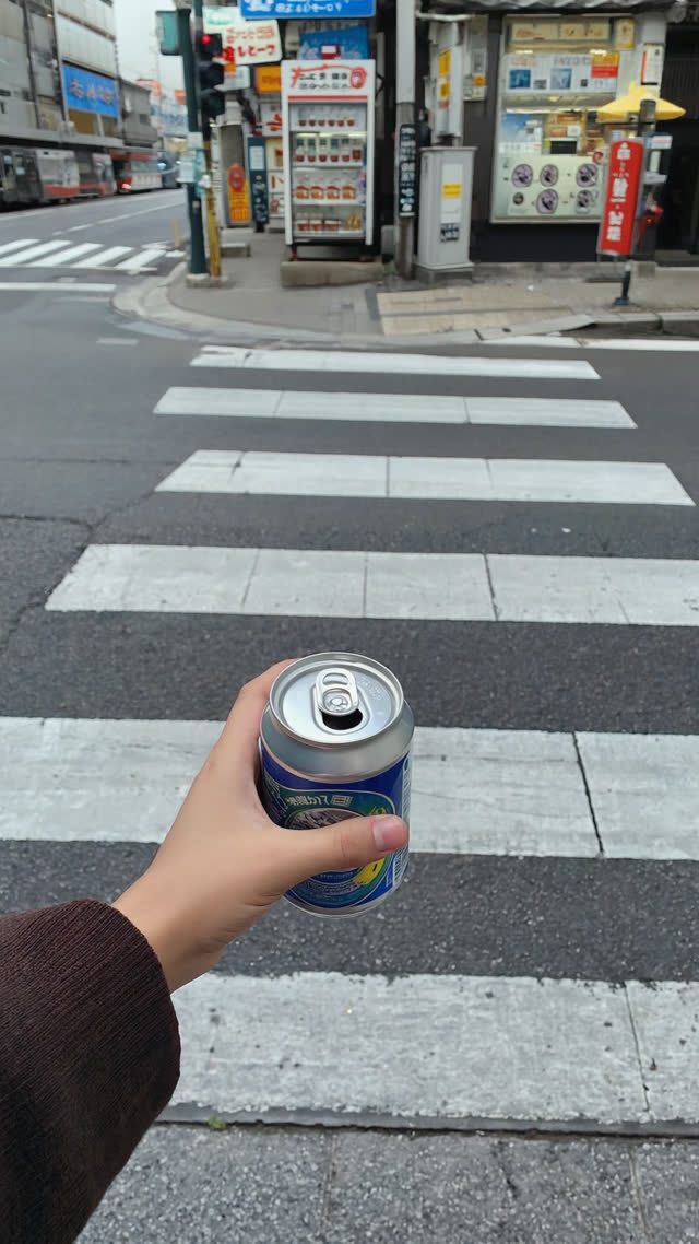 Hand holding an empty drink can on a clean Japanese street corner with no trash can anywhere, puzzled pause implied, daylight