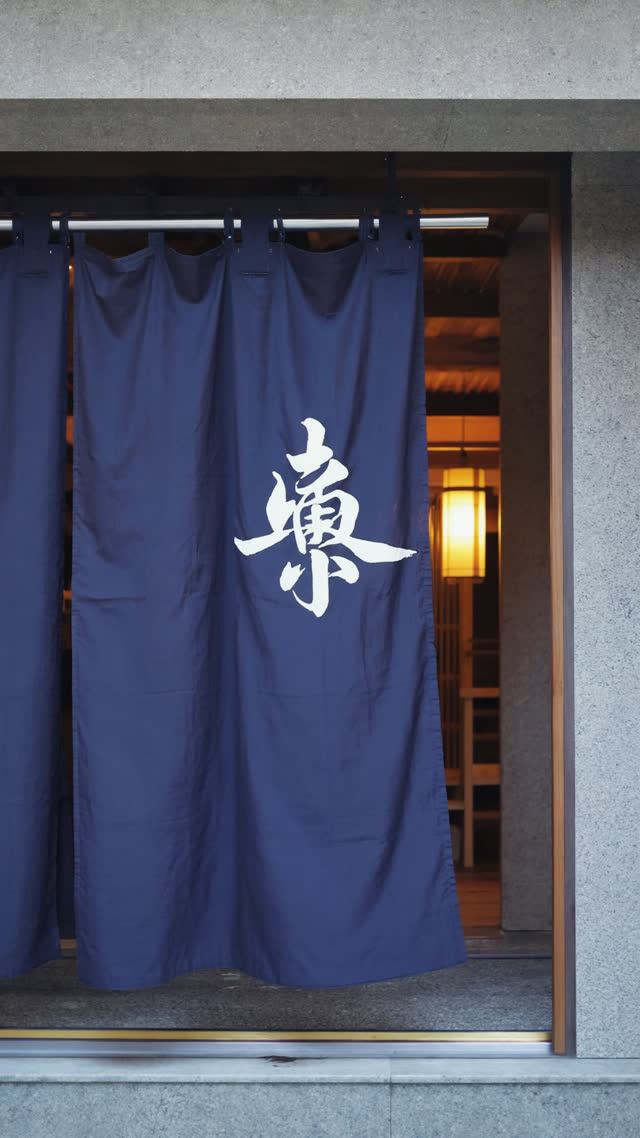 Indigo noren curtain with white Japanese character gently swaying in breeze at bathhouse entrance, fabric caught mid-motion, warm entrance light behind